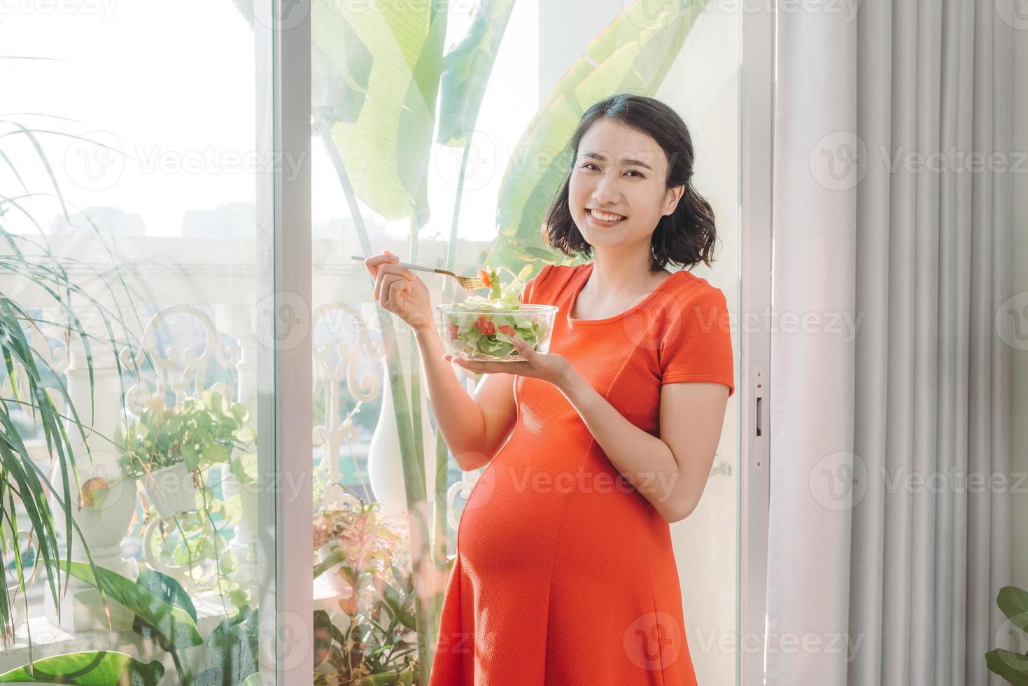 lleno de vitaminas. mujer embarazada enérgica y guapa comiendo su carne mientras lleva un plato en una mano y se relaja contra la ventana foto