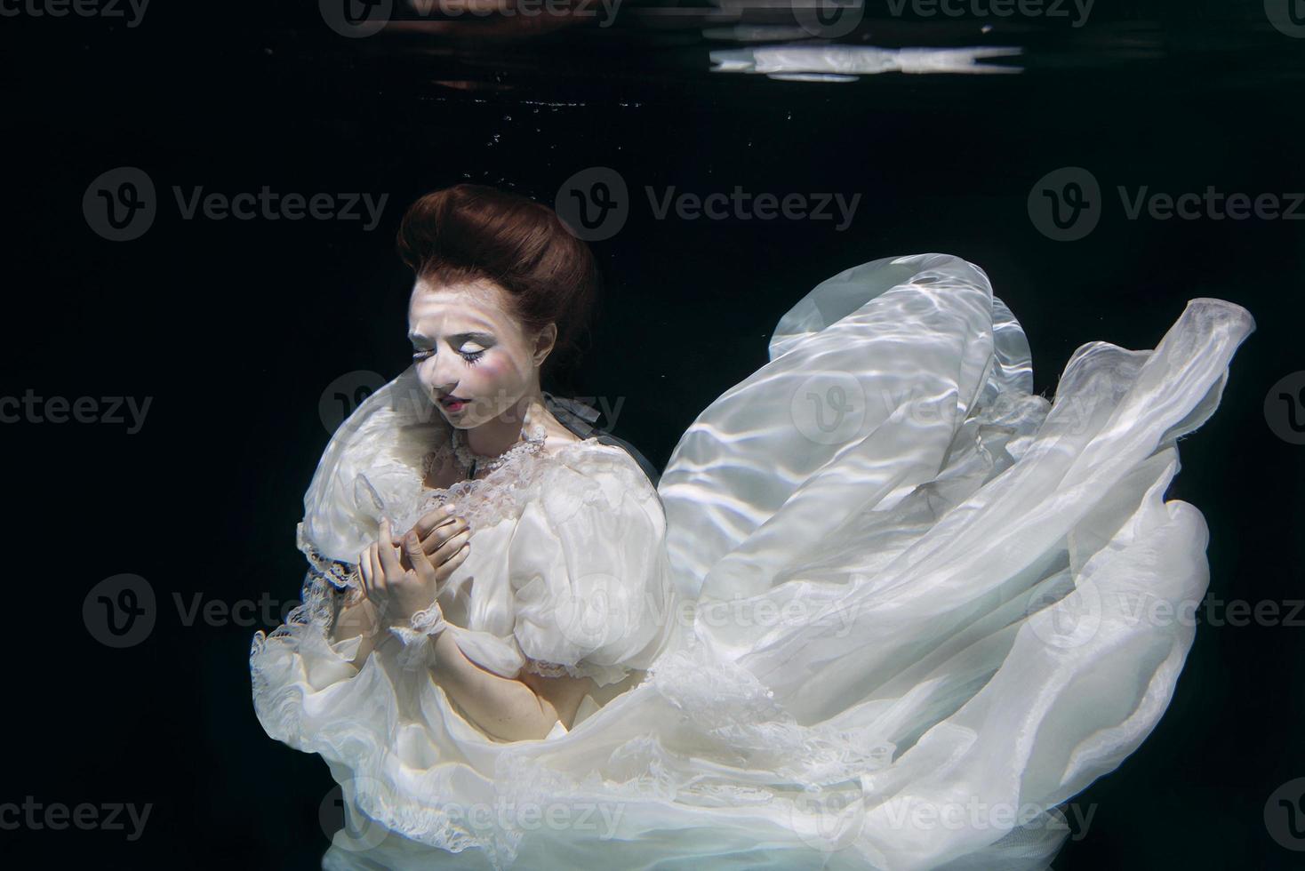 young woman in long white luxury dress underwater on the black background. Motion, art, winter concept photo