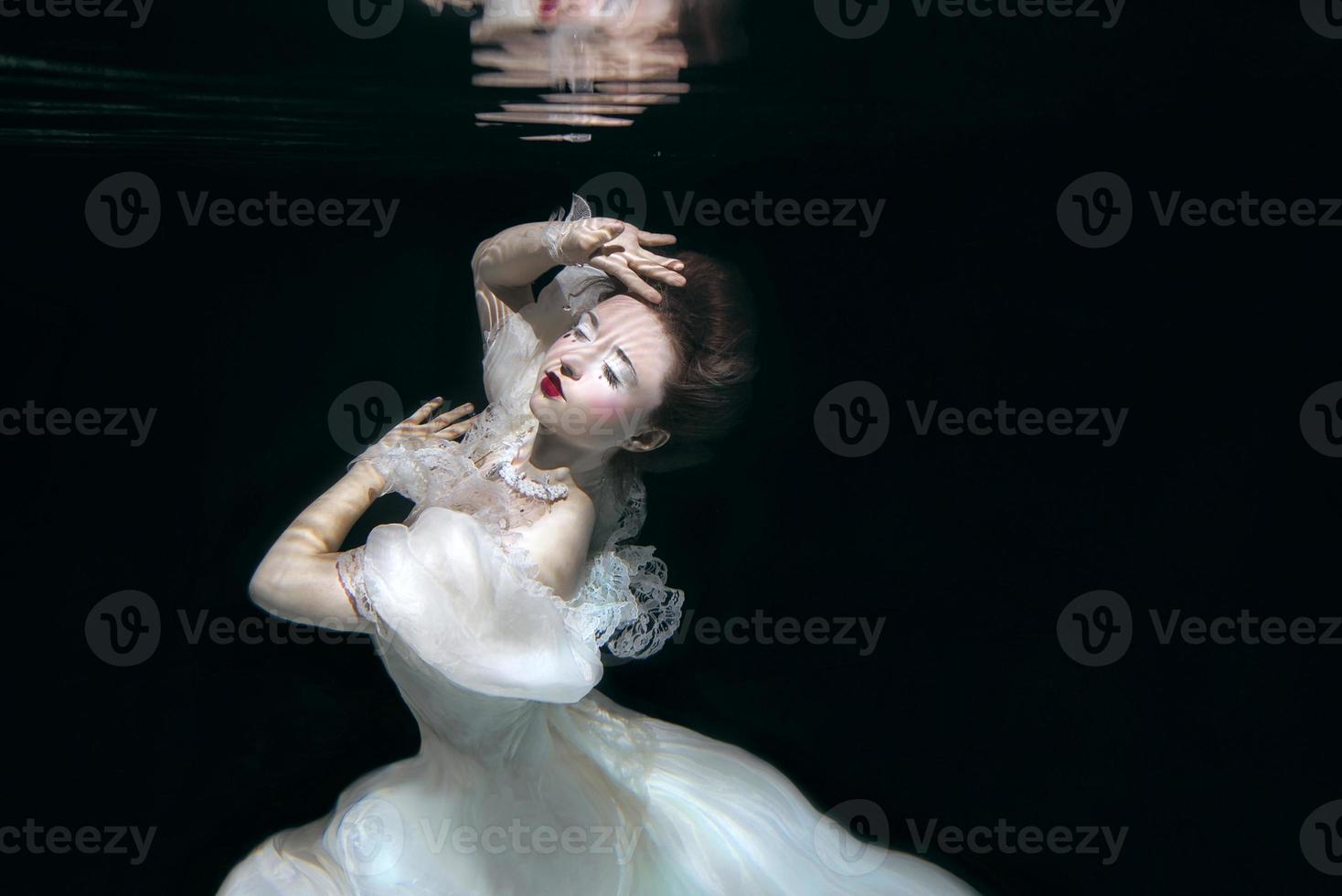 young woman in long white luxury dress underwater on the black background. Motion, art, winter concept photo