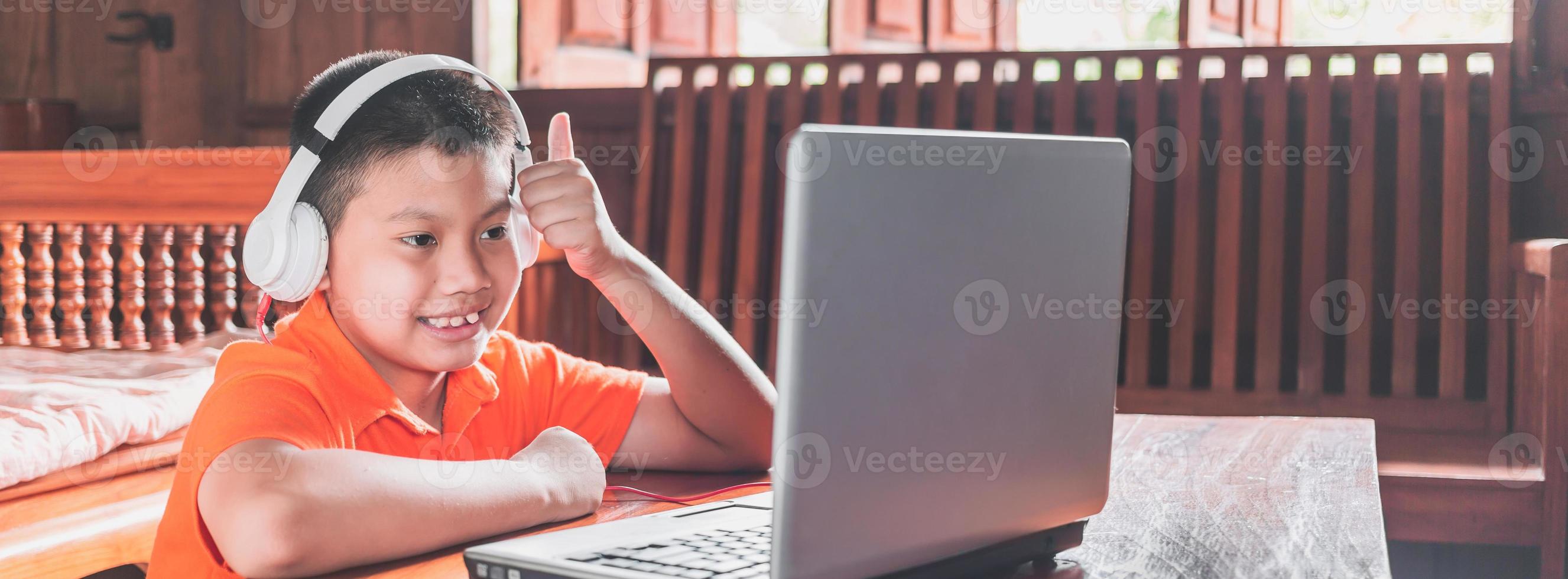 Asian child boy in headphones is using a laptop and studying online with a video call teacher at home. homeschooling, distant learning photo