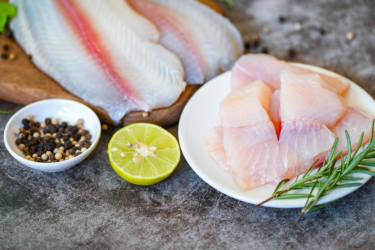 filete de pescado pangasius crudo fresco con hierbas y especias, lima limón y romero, pescado de muñeca de carne, bagre rayado de tilapia, filete de pescado en un plato blanco con ingredientes para cocinar foto