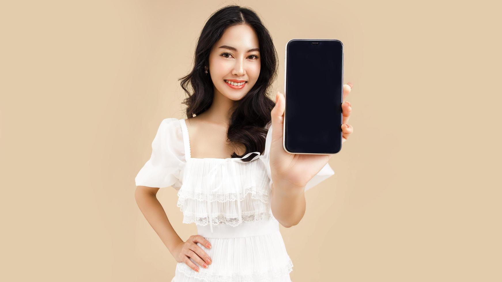 mujer asiática joven de moda que usa un teléfono inteligente de pie sobre un fondo beige aislado sintiéndose feliz. pago de compras en línea con teléfono móvil. mujer mostrando teléfono celular de pantalla en blanco. foto