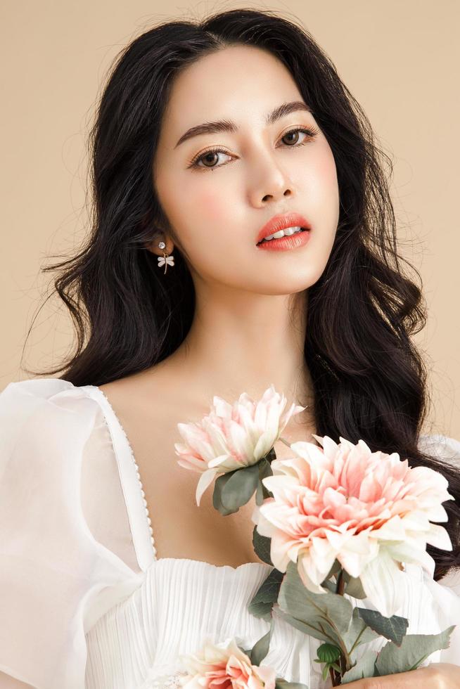 mujer asiática con una cara hermosa y una piel limpia y fresca perfecta con flores. lindo modelo femenino con maquillaje natural y ojos brillantes sobre fondo beige aislado. foto