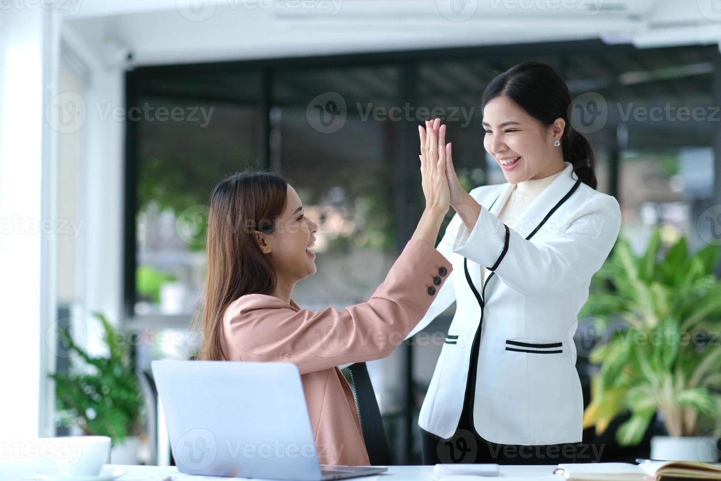 Two young Asian businesswomen show joyful expression of success at work smiling happily with a laptop computer in a modern office. photo