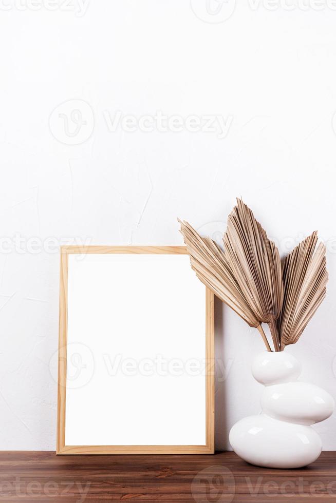 White frame mockup with dry palm leaves in a vase on wooden table photo