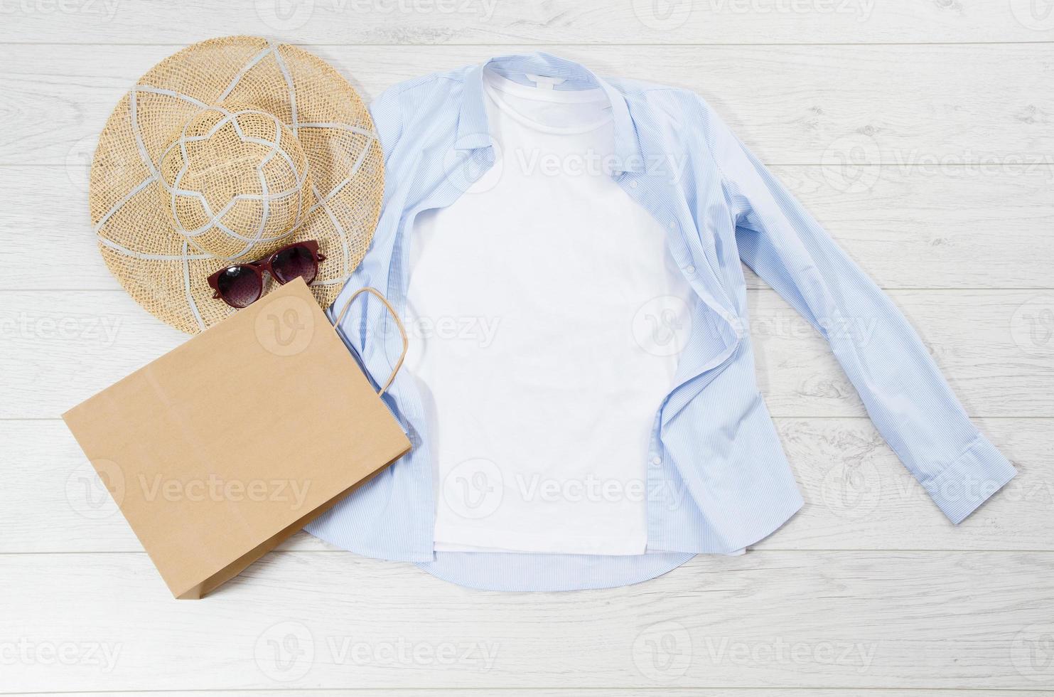 White closeup t shirt mock up flat lay on wooden background. Summer accessories. Hat, sunglasses and paper bag. Top view and copy space. Mockup t-shirt and summertime. Template blank shirt. photo