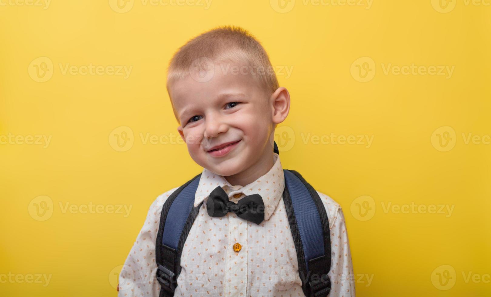 De vuelta a la escuela. un chico rubio y divertido con una mochila en un pantalones claro sobre un fondo amarillo sonríe y mira a la cámara. copie el espacio foto