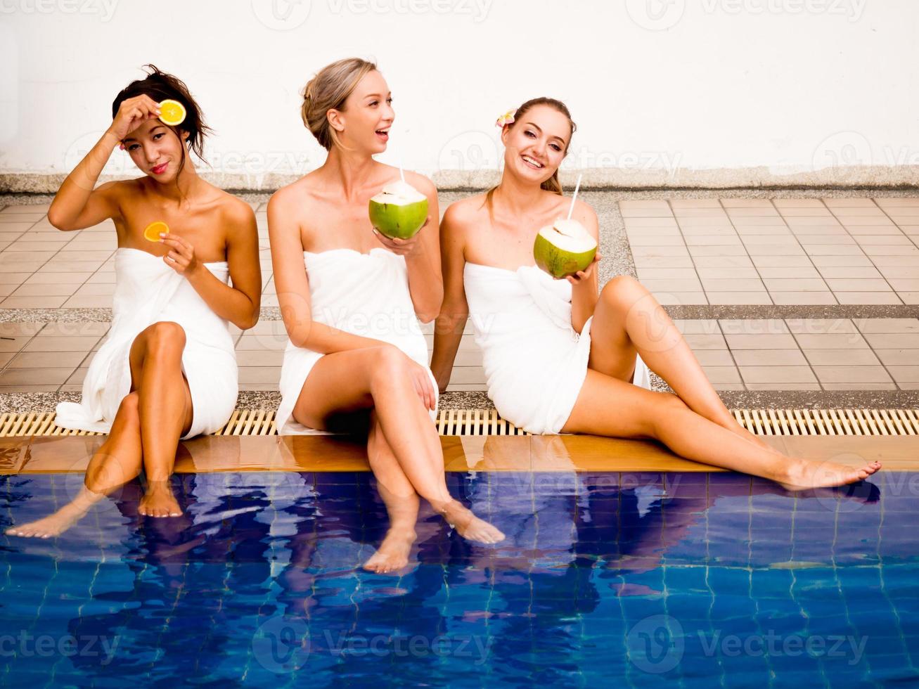 Beautiful woman wrapped with towel.petty girls holding coconut and slide orange.happy female talking together near swimming pool. photo