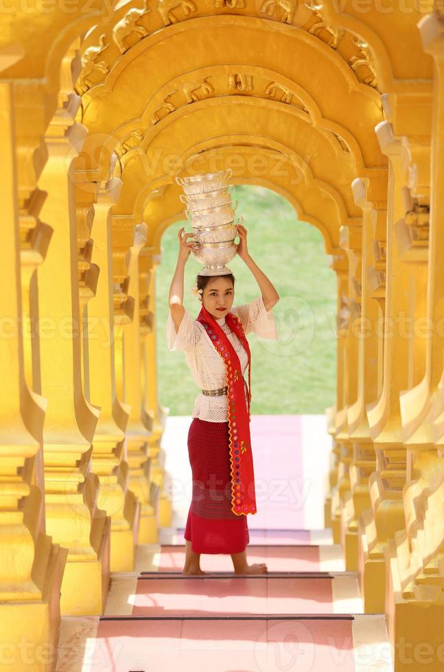 joven asiática con traje tradicional birmano sosteniendo un tazón de arroz a mano en la pagoda dorada en el templo de myanmar. mujeres de myanmar sosteniendo flores con vestido tradicional birmano visitando un templo budista foto