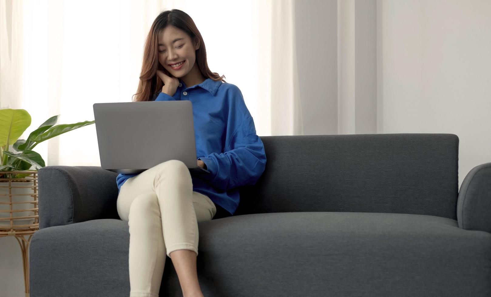 joven mujer asiática independiente de negocios que trabaja en una computadora portátil revisando las redes sociales mientras está acostada en el sofá cuando se relaja en la sala de estar en casa. foto