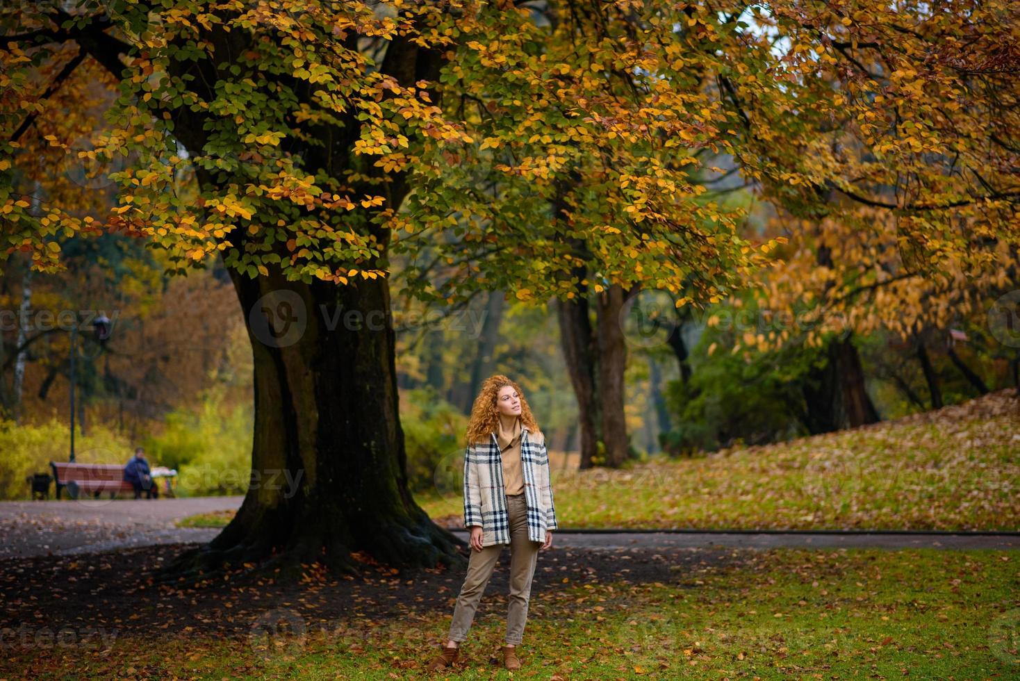 Beautiful red-haired girl with curly hair and blue eyes. The girl is wearing a checkered jacket. The girl is sitting on a bench. photo