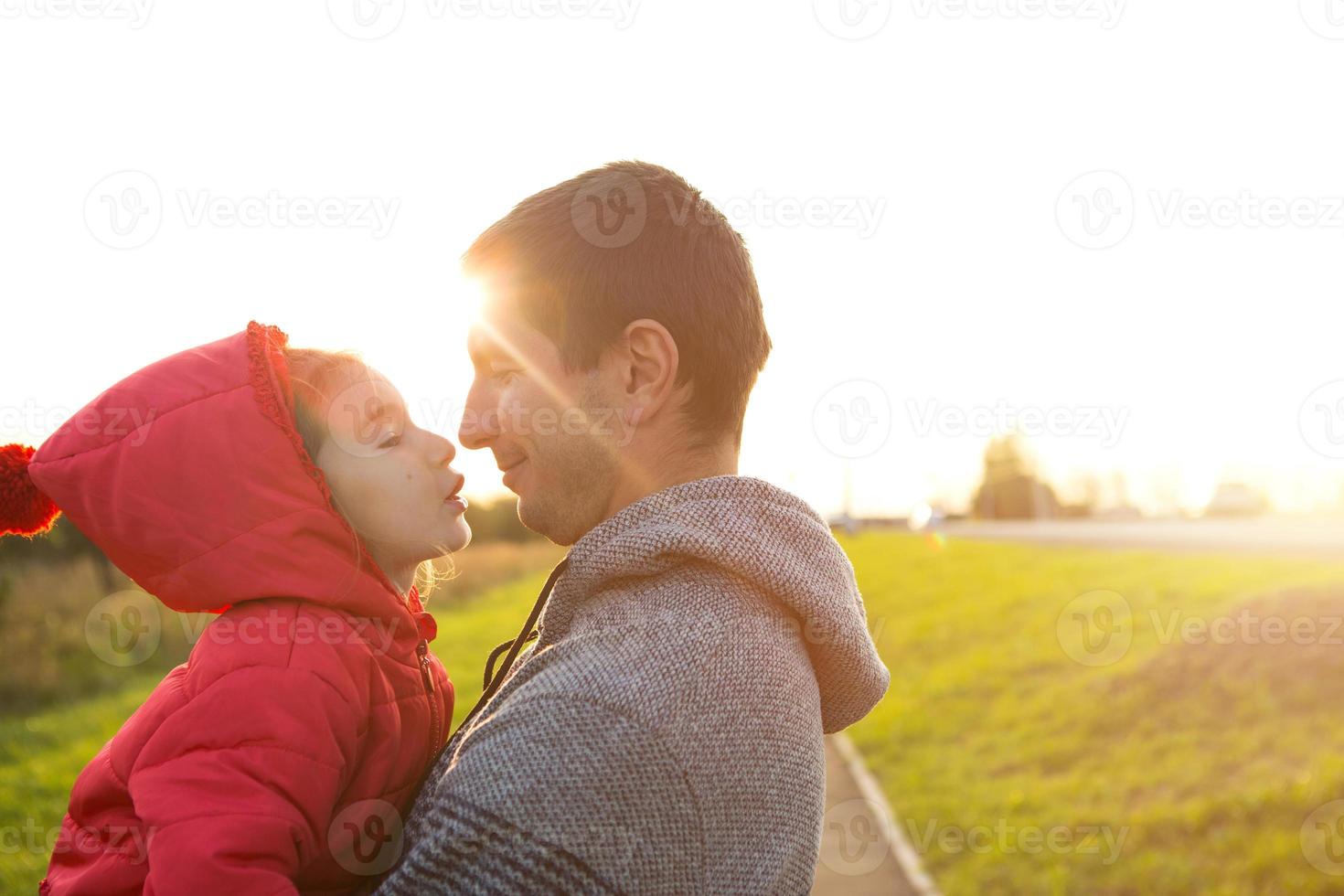 Little girl in red jacket with a hood hugs and kisses her dad, smiles, touches her nose. Happy family, children's emotions, father's day, bright rays of the sun, Caucasian appearance. Space for text. photo
