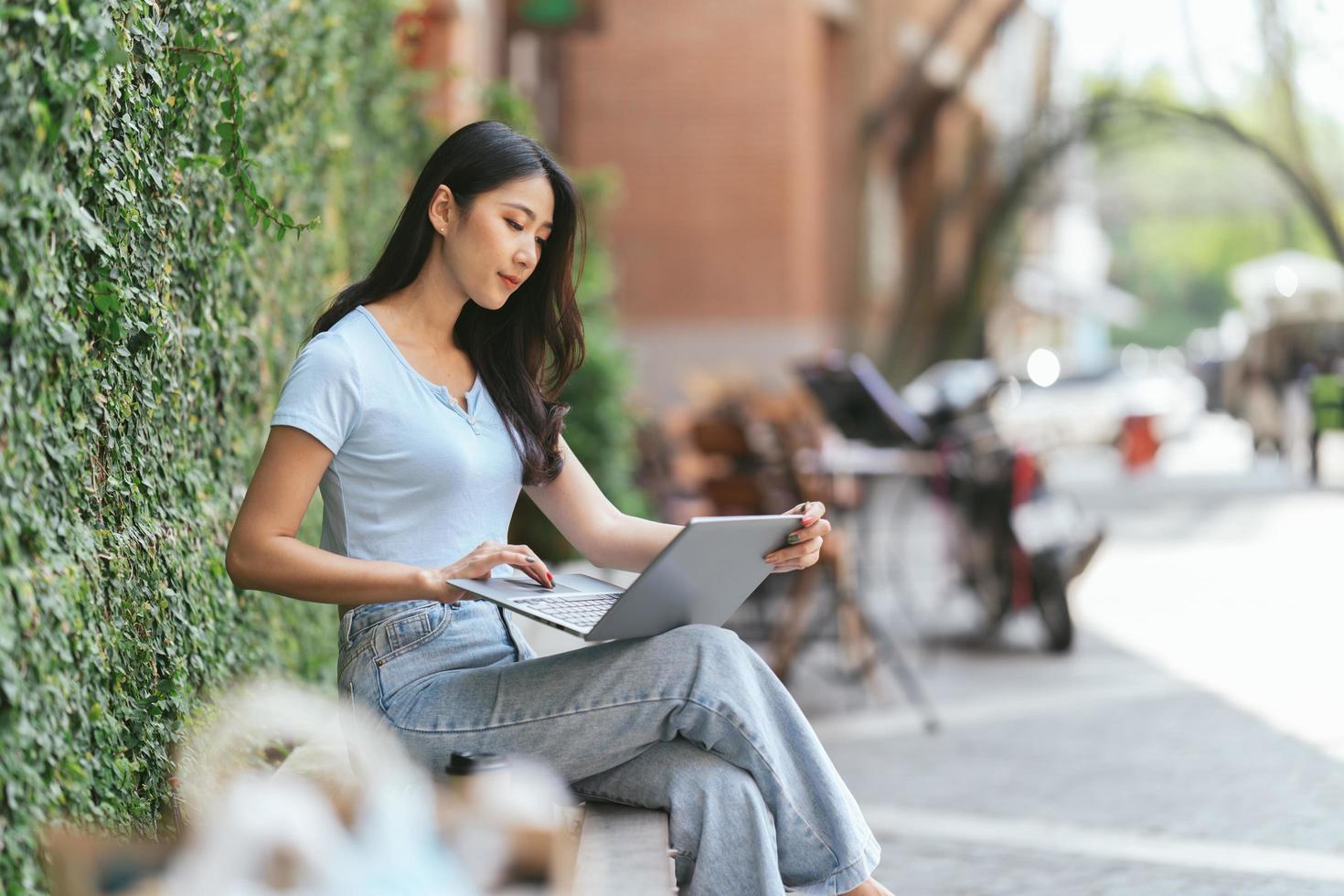 Portrait of beautiful Asian woman sitting outdoors at coffee shop restaurant during summer, using smart wireless technology computer laptop and smartphone, relaxing coffee break at cafe restaurant. photo