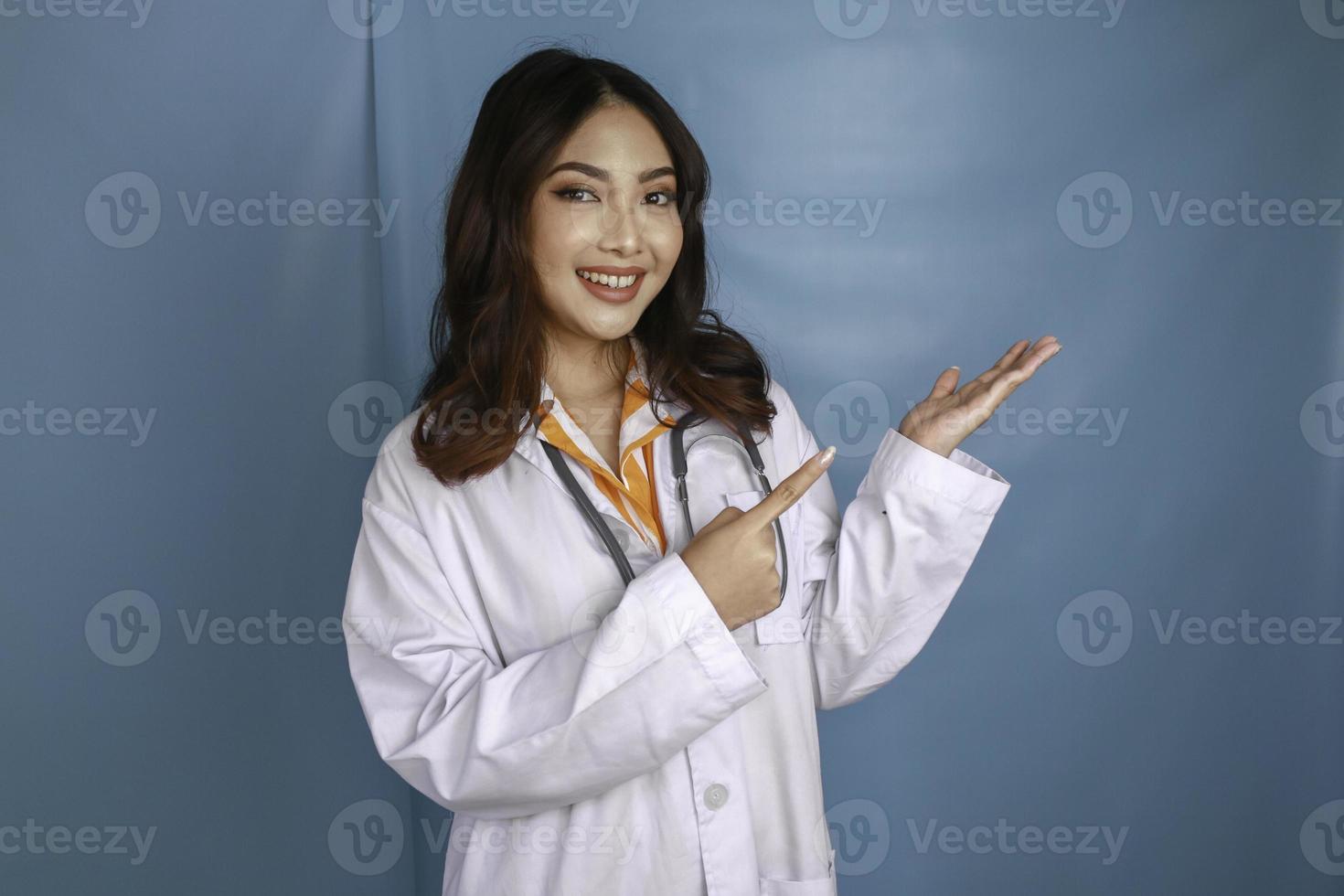 Portrait of a young Asian woman doctor, medical professional is smiling and pointing at a copy space isolated over blue background photo