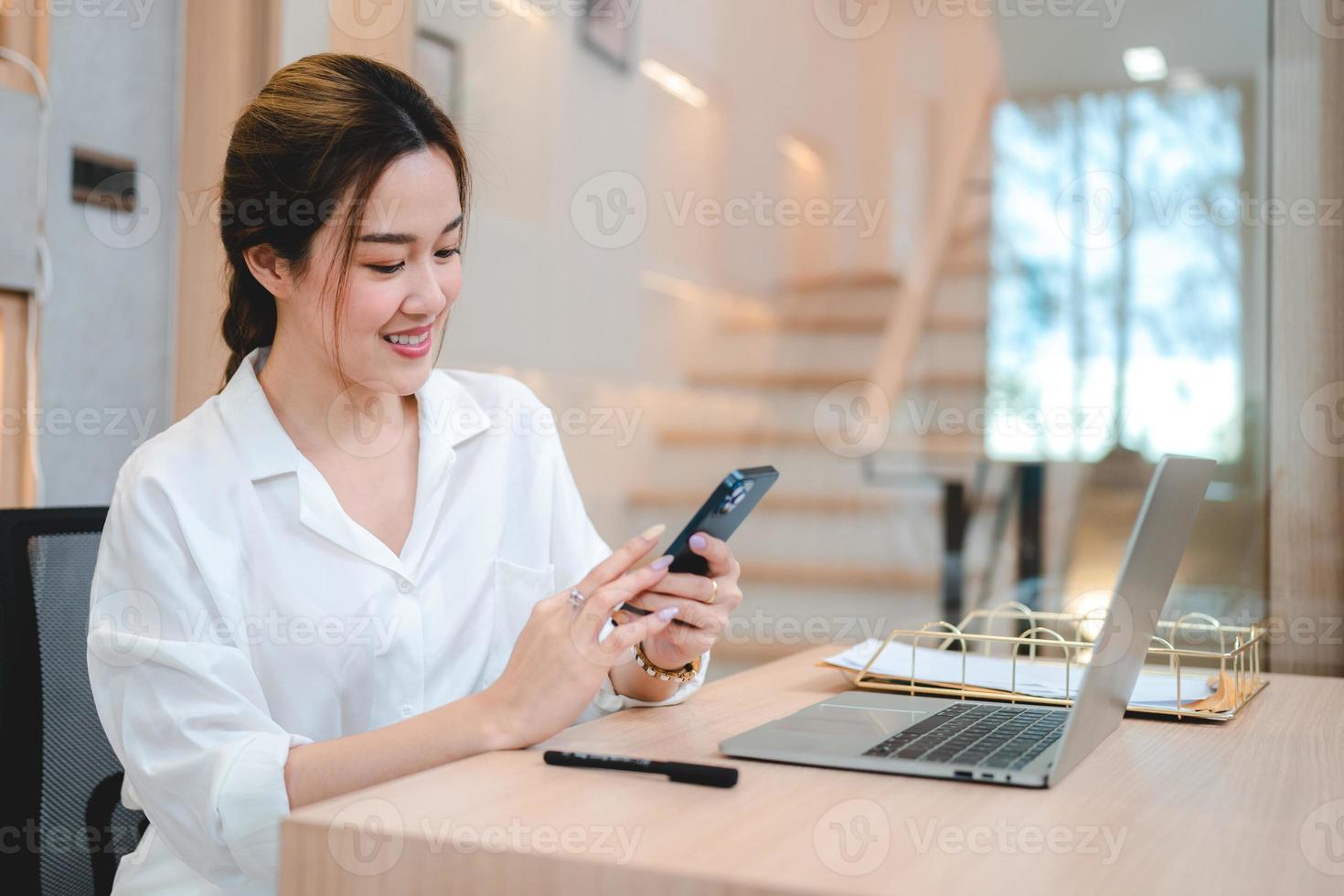 Smiling young Asian business woman and entrepreneur using mobile phone and scrolling and browsing through social media on taking break after working on laptop photo