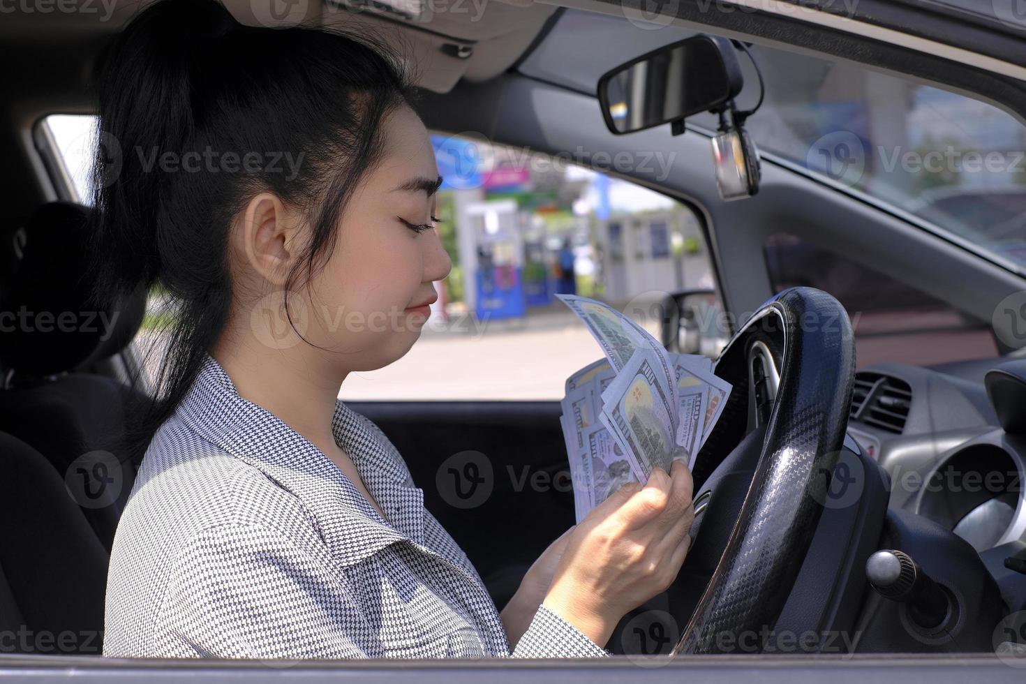 Closeup portrait of a happy smiling attractive Asian woman holds cash dollar bills sitting inside her car at gas station photo