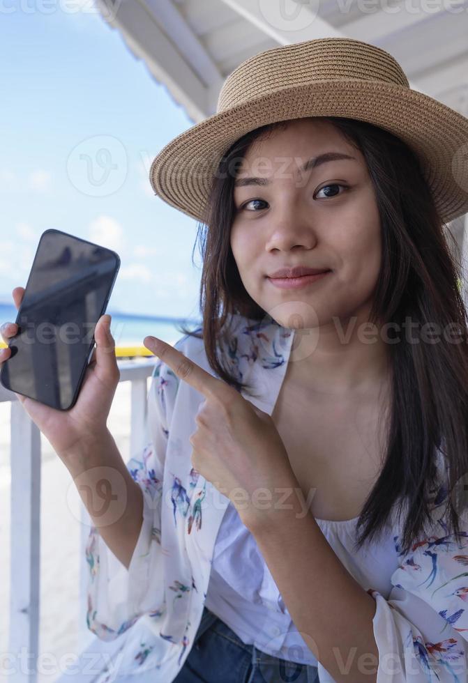 Traveler girl showing smartphone blank screen advertising application to shopping and paid online by mobile app. Young Asia lady show empty smartphone screen with a positive expression. photo