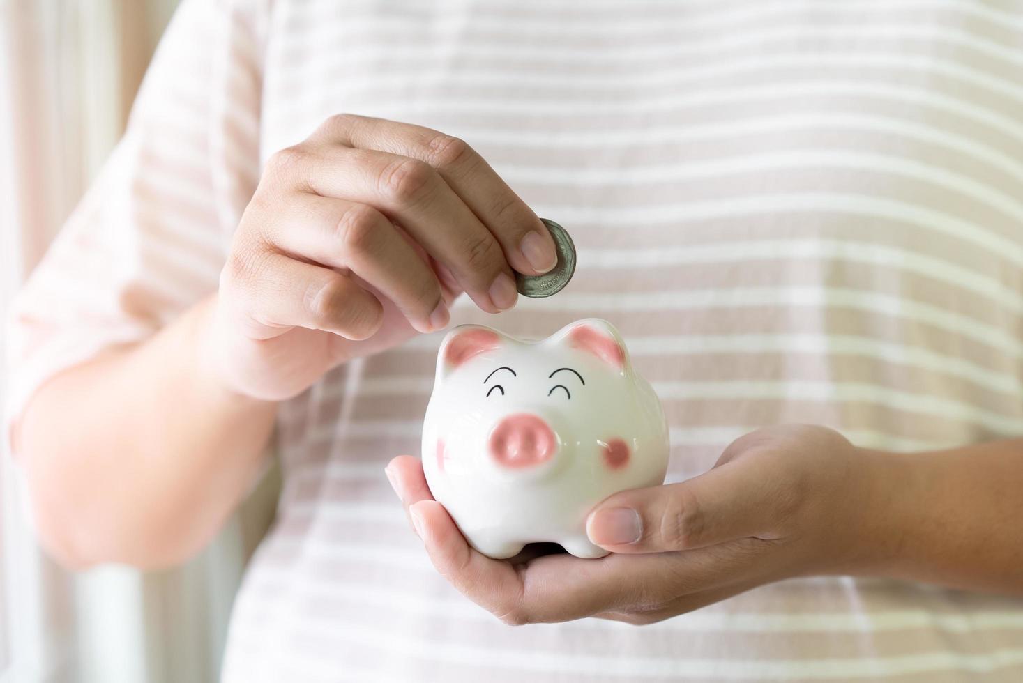 Woman hands holding and putting Thai currency coin into piggy bank. Saving concept with piggy bank indoor with sun light. photo