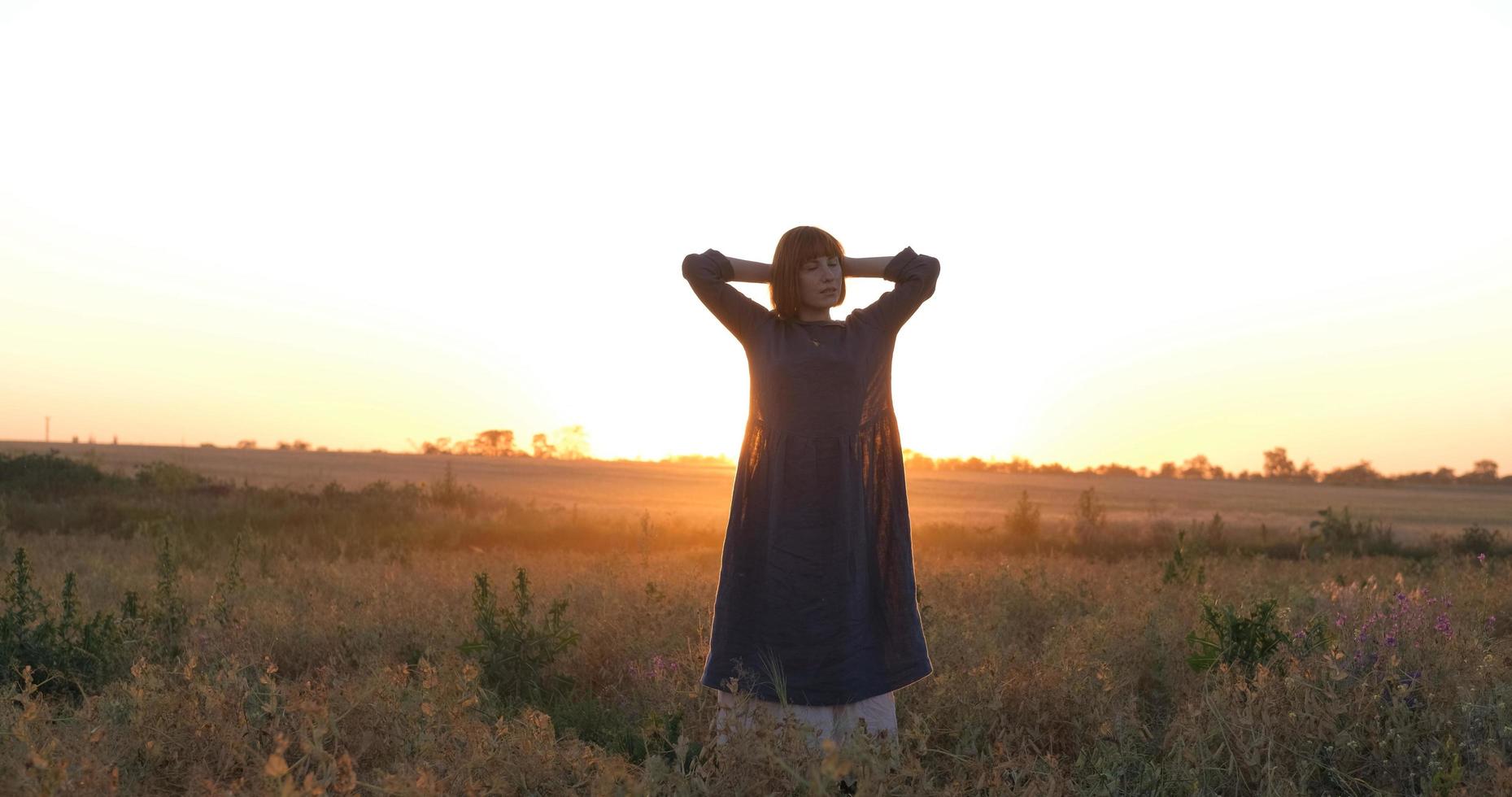 joven pelirroja con hermoso vestido boho relajándose en el campo durante la puesta de sol nublada, mujer al aire libre con ramo en las manos foto