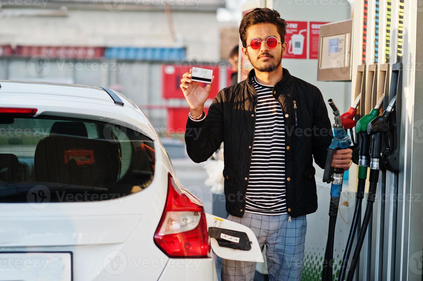 South asian man or indian male refueling his white car on gas station and hold credit card. photo
