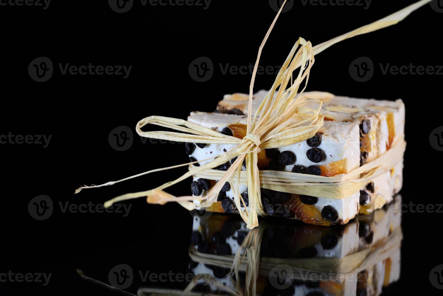 Several pieces of torrone or white nougat with nuts, candied fruits and chocolate are tied together with raffia and lie on a reflective surface in front of a dark background photo