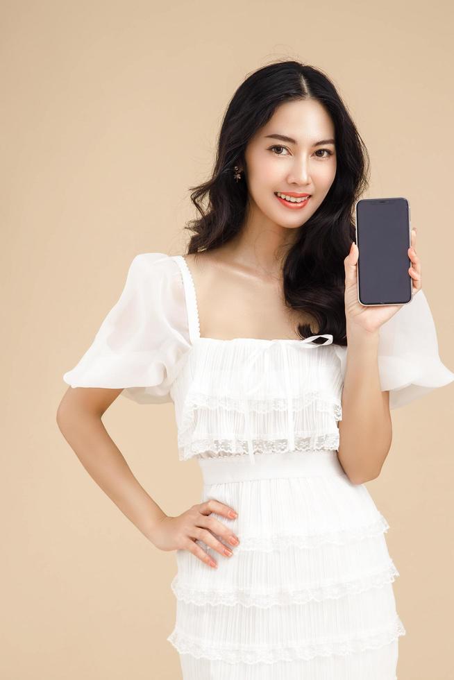mujer asiática joven de moda que usa un teléfono inteligente de pie sobre un fondo beige aislado sintiéndose feliz. pago de compras en línea con teléfono móvil. mujer mostrando teléfono celular de pantalla en blanco. foto