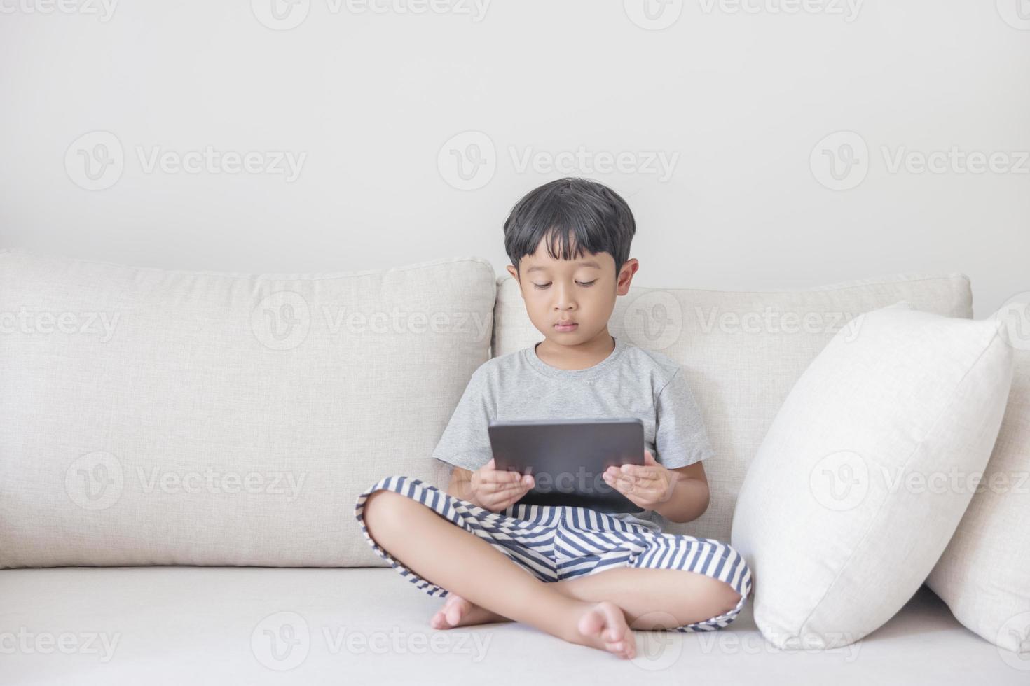 Adorable happy Asian boy wearing a gray shirt and blue-white striped shorts is having fun playing with his tablet on a cream sofa. looking at the mobile screen photo