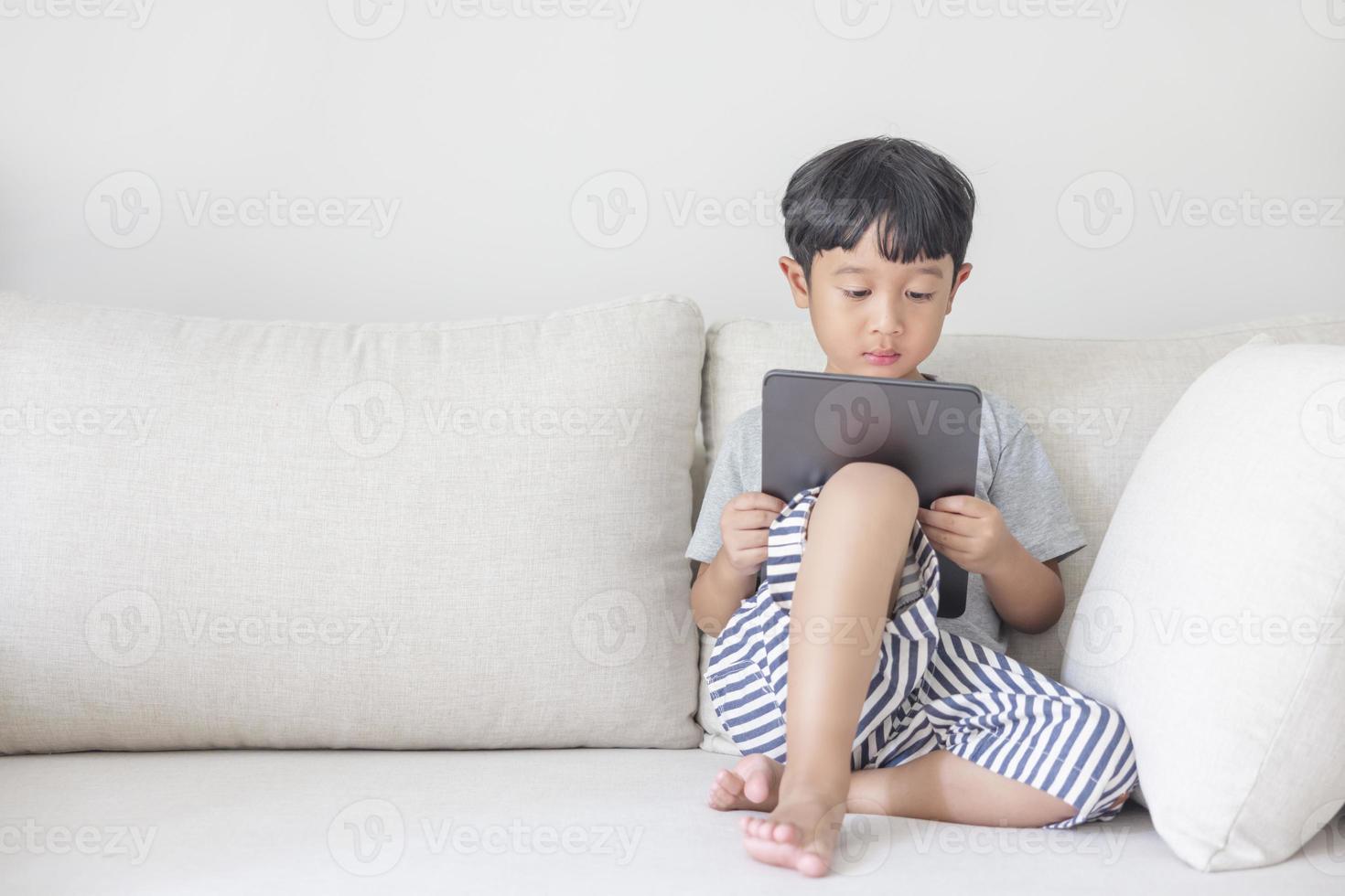 Adorable happy Asian boy wearing a gray shirt and blue-white striped shorts is having fun playing with his tablet on a cream sofa. looking at the mobile screen photo