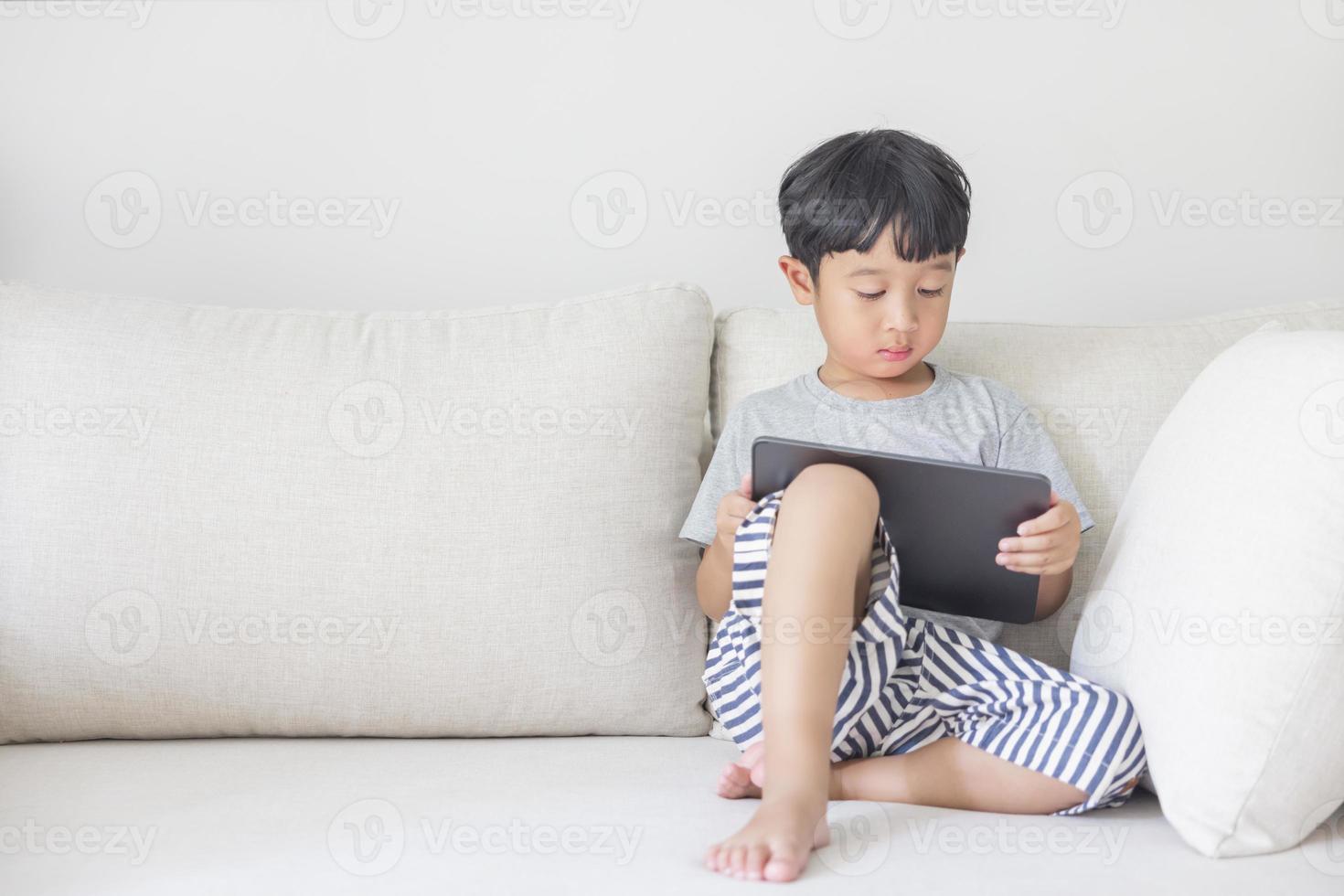 adorable niño asiático feliz con una camisa gris y pantalones cortos de rayas azules y blancas se divierte jugando con su tableta en un sofá color crema. mirando la pantalla del movil foto