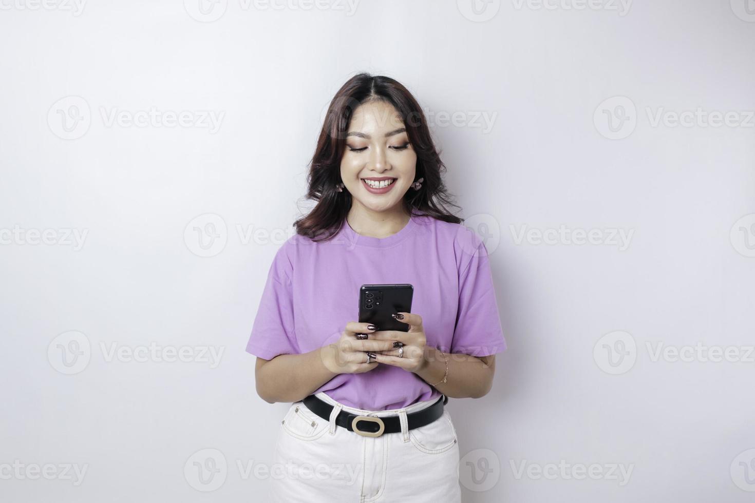 A portrait of a happy Asian woman is smiling and holding her smartphone wearing a lilac purple t-shirt isolated by a white background photo