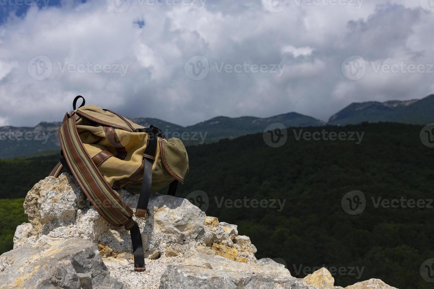 A brown-green backpack lies on a large gray stone on a rock against the backdrop of green mountains. photo
