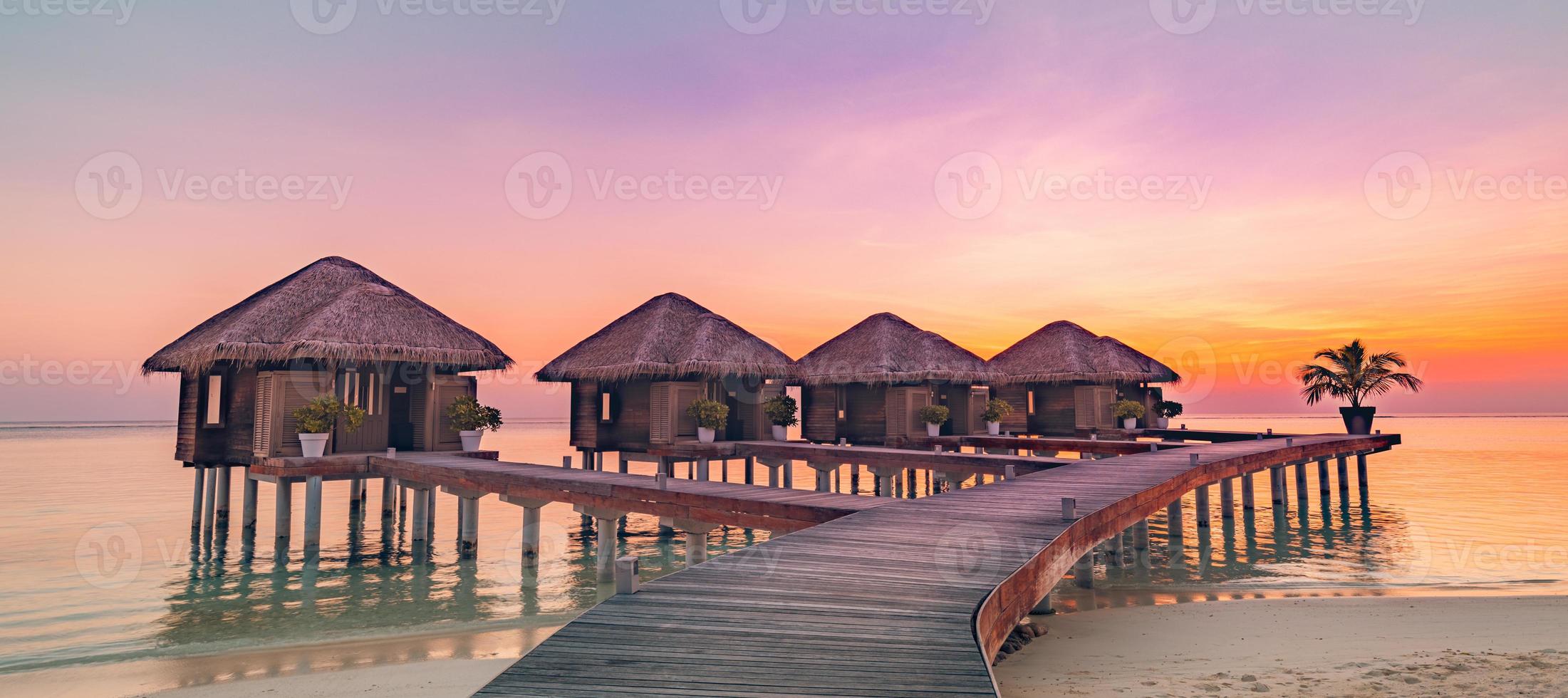 Maldives island sunset panorama. Water bungalows resort at islands beach. Indian Ocean, Maldives. Beautiful sunset landscape, luxury resort villas and colorful sky. Summer vacation holiday travel photo