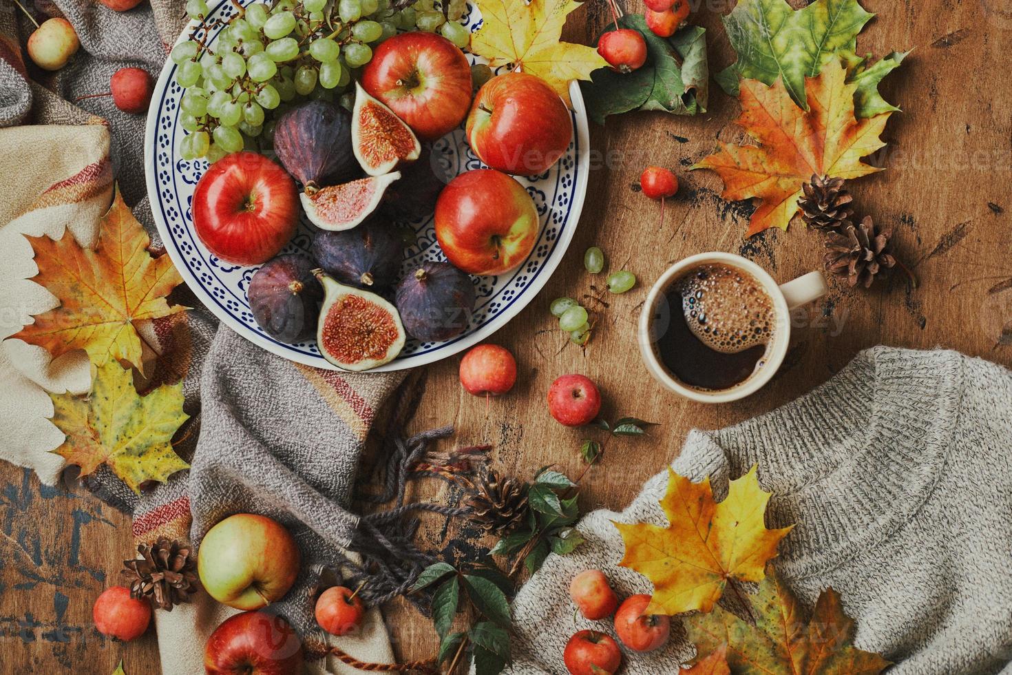 Dish with figs, apples and grapes and cup of coffee on wooden background with a warm sweater, scarf, autumn leaves and apples. Autumn background, top view. photo