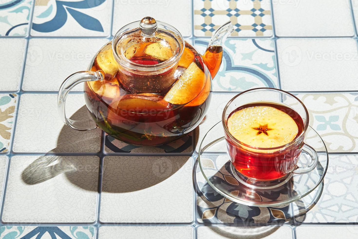 té de frutas con manzanas y tomillo en una tetera de vidrio y una taza sobre una mesa hecha de azulejos de colores foto