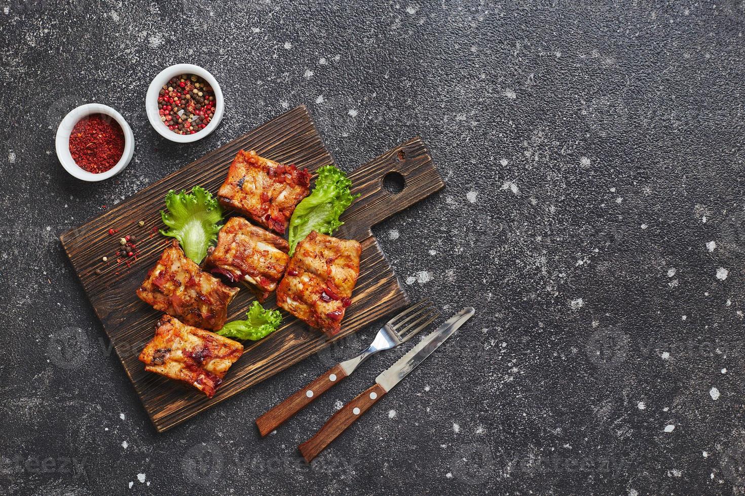 Grilled baked pork ribs with spices and vegetables on wooden cutting board on dark background. American food concept. Top view, copy space. photo