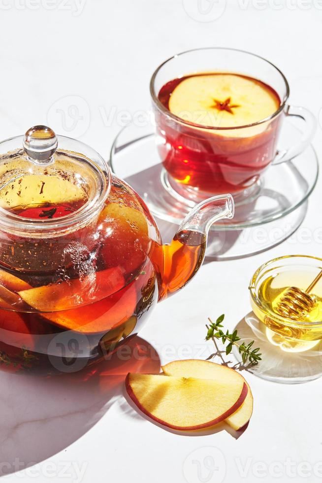 té de frutas con manzanas y tomillo y miel en tetera de vidrio y taza sobre fondo blanco con sombras duras foto
