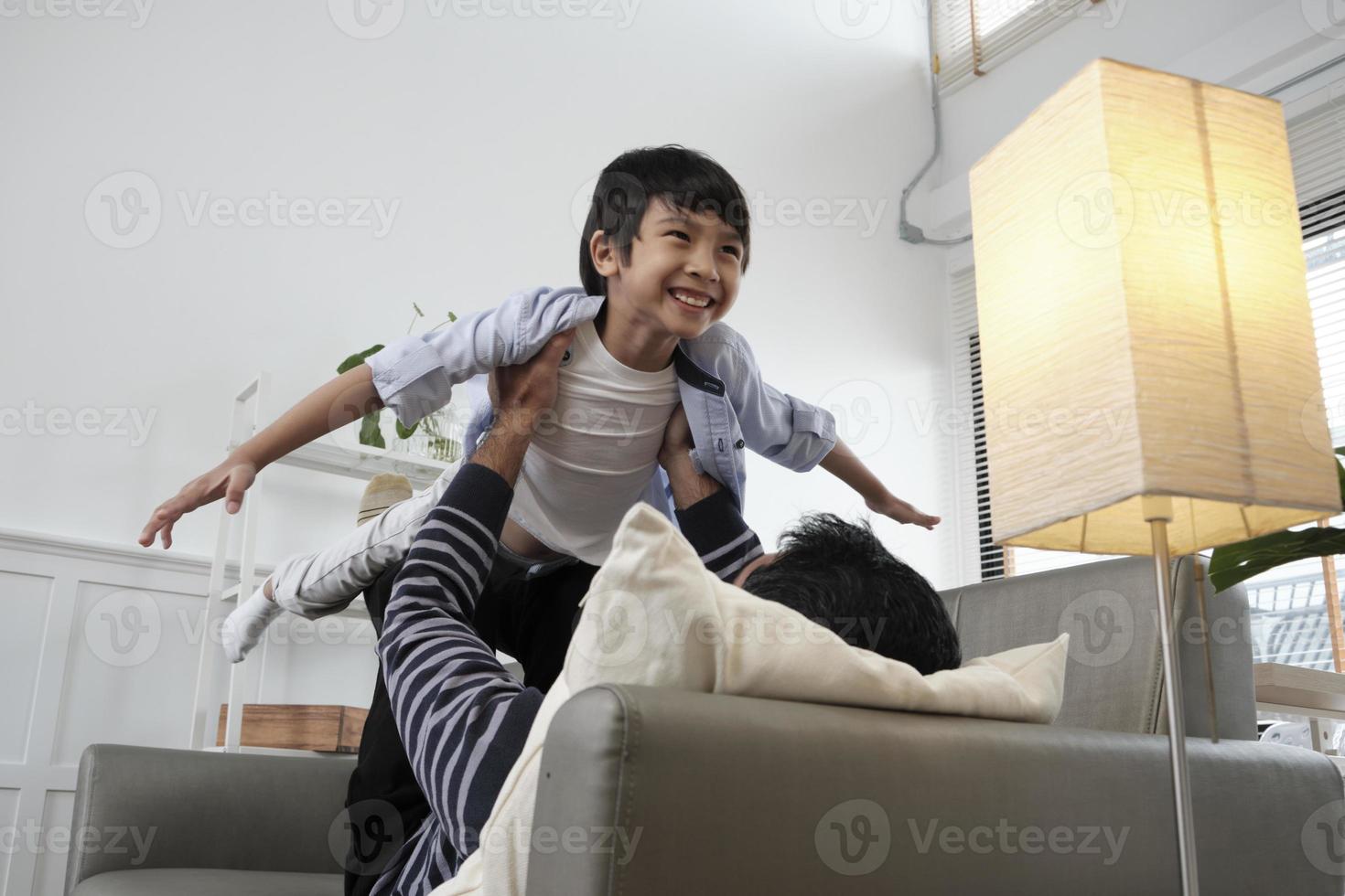 Asian Thai family together, father lies down and fun plays with son by lifting, spread arms like flying airplane on living room sofa, happy leisure times, lovely weekend, wellbeing domestic lifestyle. photo