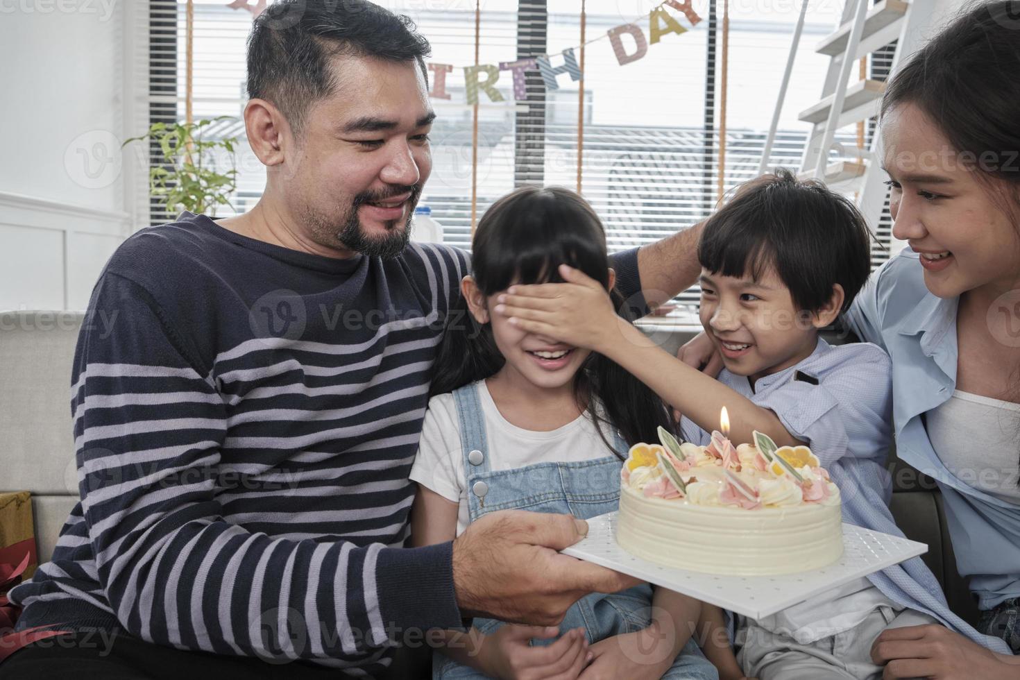 feliz familia tailandesa asiática, la joven hija se sorprende con un pastel de cumpleaños, sopla una vela, reza y celebra alegremente la fiesta con los padres juntos en la sala de estar, el bienestar del estilo de vida del evento doméstico. foto