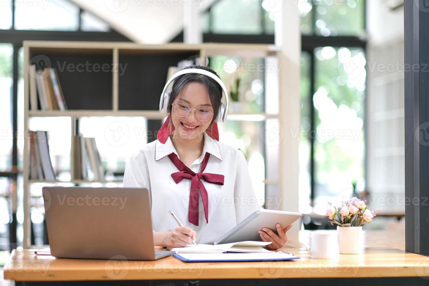 Young adult happy smiling Hispanic Asian student wearing headphones talking on online chat meeting using laptop in university campus or at virtual office. College female student learning remotely. photo