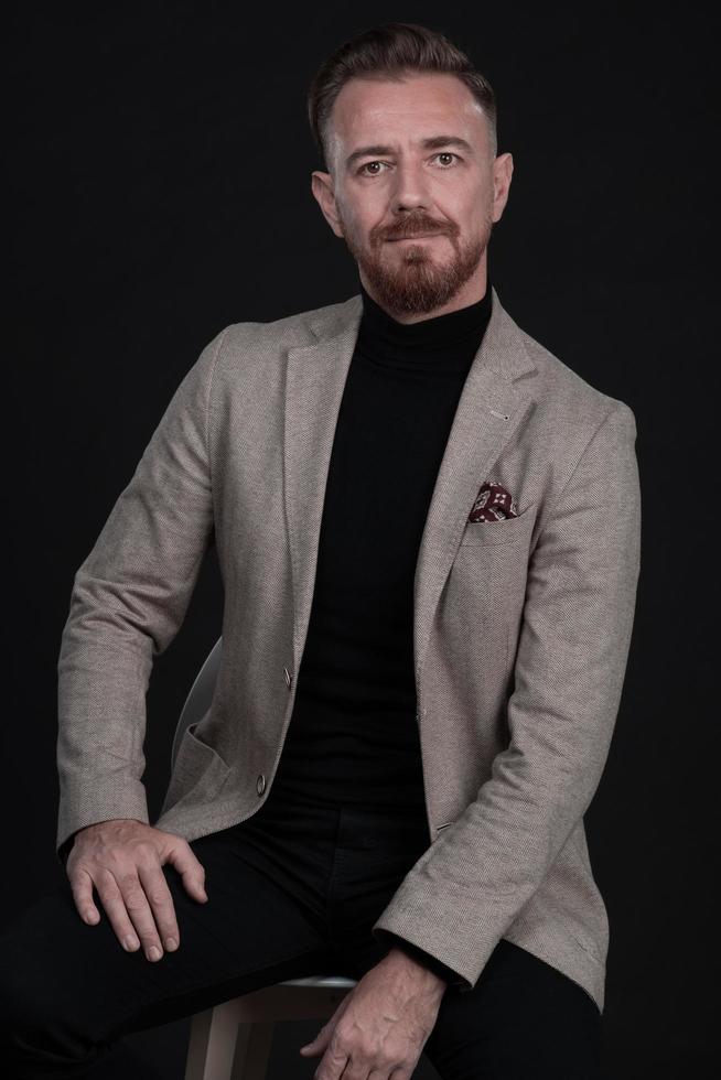 Portrait of adult businessman wearing trendy suit and sitting in modern studio on stylish chair against the black background photo