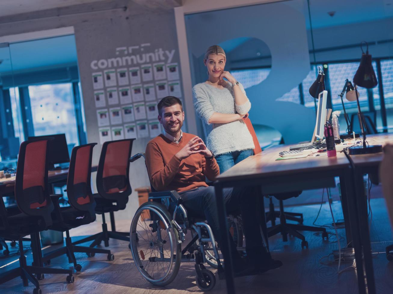 Businessman in a wheelchair in modern coworking office space working late night in office. Colleagues in background. Disability and handicap concept. photo