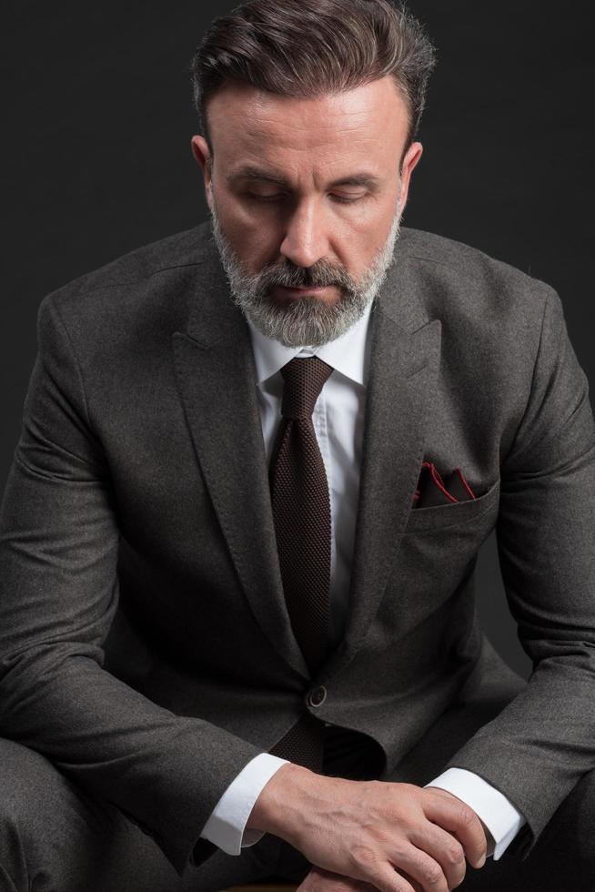 Portrait of adult businessman wearing trendy suit and sitting in modern studio on stylish chair against the black background photo