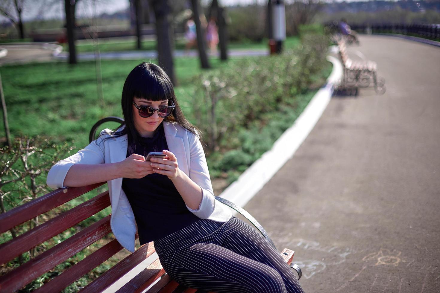 Cute Woman with Sunglasses and Long Hair is Using Mobile Device Enjoying Sunbeams and Warm Day Outdoors. Portrait of Smiling Brunette with Smartphone in the Park. photo
