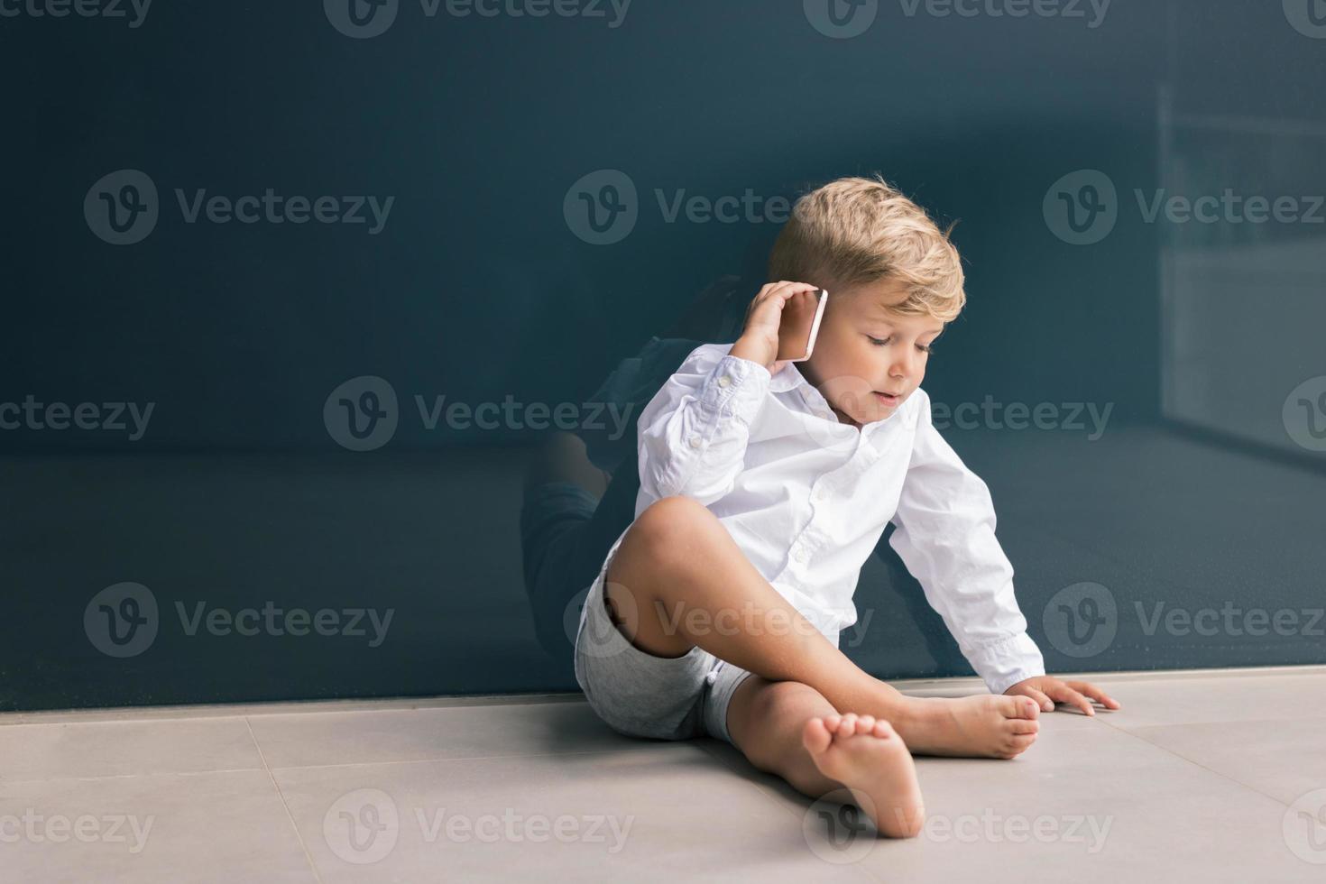 niño hablando por teléfono móvil mientras se relaja en el suelo contra la pared. foto