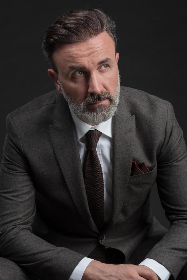 Portrait of adult businessman wearing trendy suit and sitting in modern studio on stylish chair against the black background photo