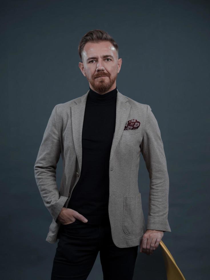 Portrait of adult businessman wearing trendy suit and sitting in modern studio on stylish chair against the black background photo