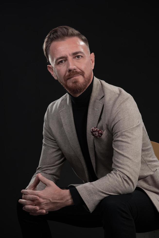 Portrait of adult businessman wearing trendy suit and sitting in modern studio on stylish chair against the black background photo