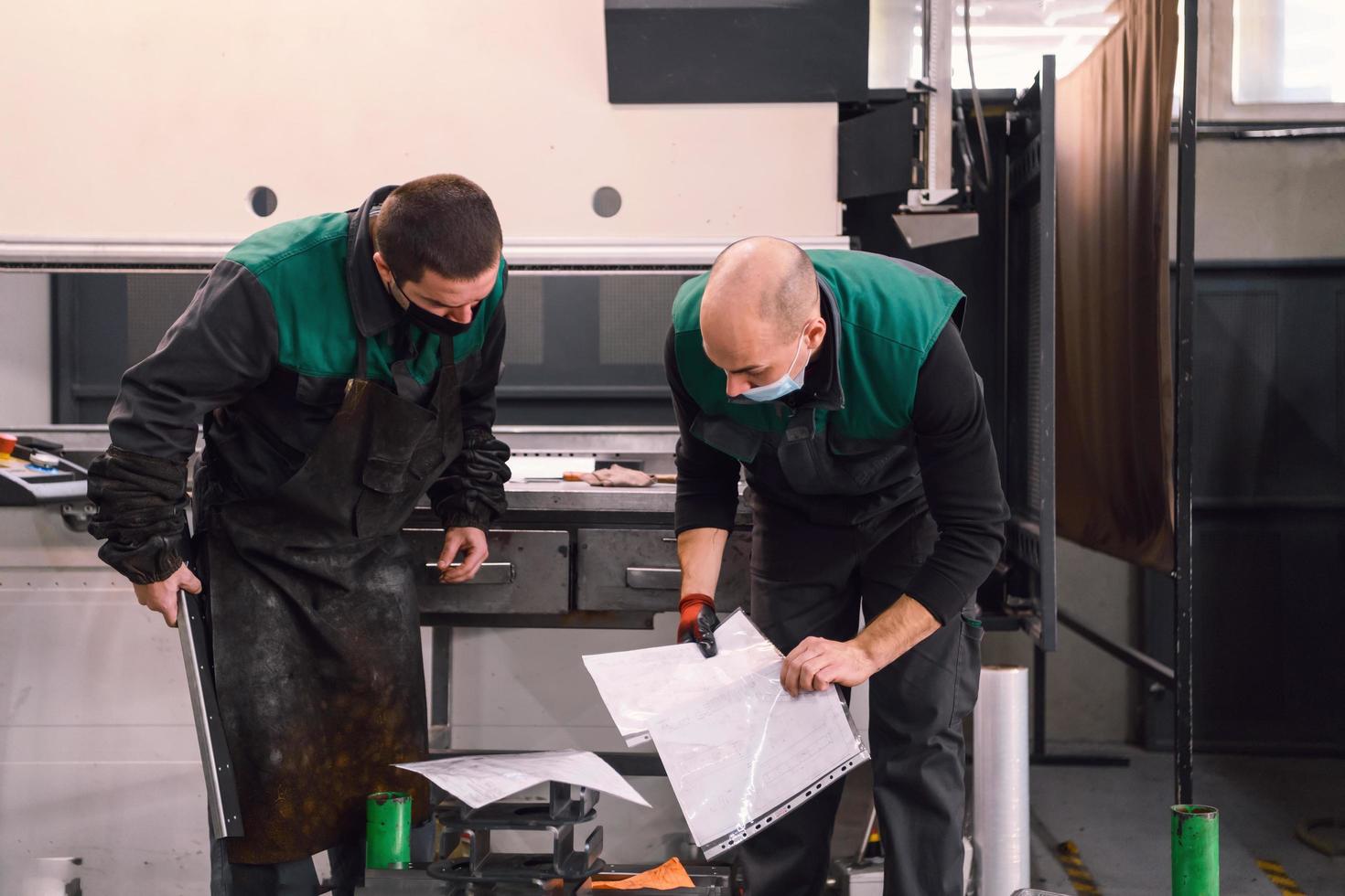 dos trabajadores que usan una máscara protectora debido a una pandemia de coronavirus, trabajan en una fábrica moderna y preparan un programa y material para una máquina cnc foto