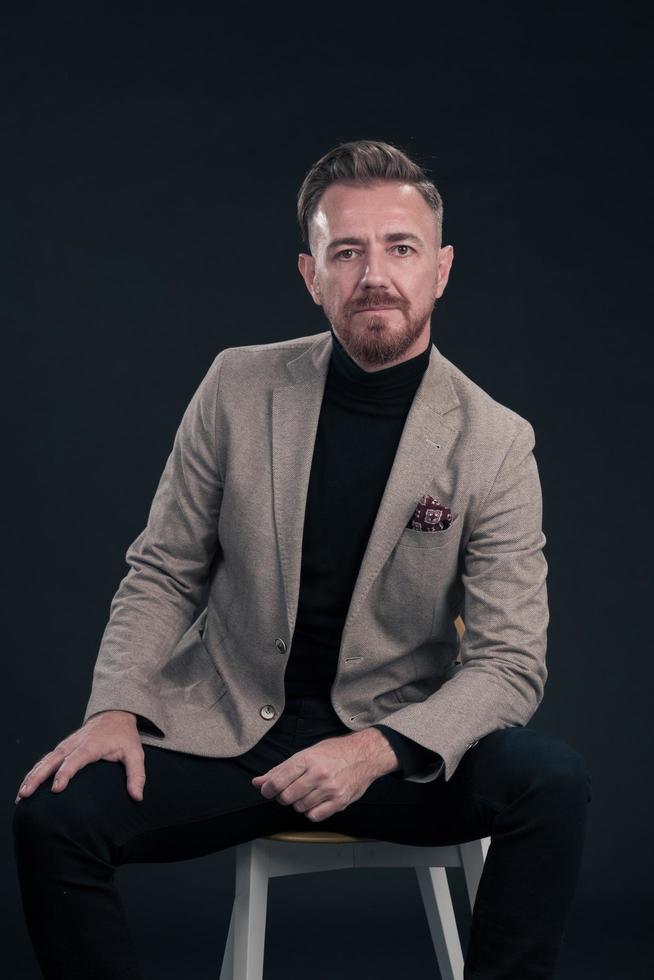 Portrait of adult businessman wearing trendy suit and sitting in modern studio on stylish chair against the black background photo