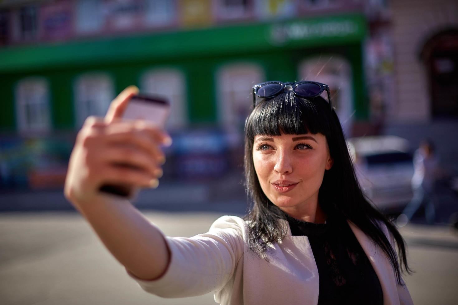 Cute Woman with Sunglasses and Long Hair is Using Mobile Device Enjoying Sunbeams and Warm Day Outdoors. Portrait of Smiling Brunette doing selfie on a smartphone photo